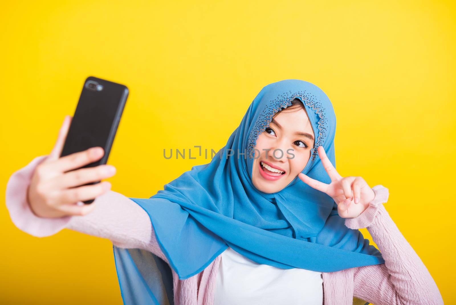 Asian Muslim Arab, Portrait of happy beautiful young woman Islam religious wear veil hijab funny smile she taking making selfie with smart mobile phone, studio shot isolated on yellow background