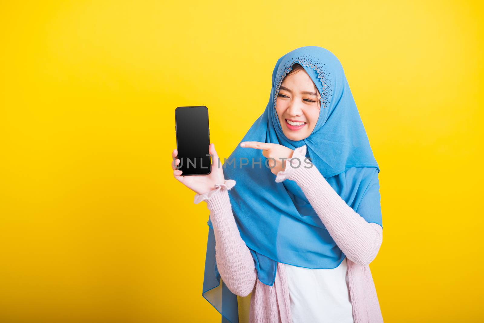 Asian Muslim Arab, Portrait of happy beautiful young woman Islam religious wear veil hijab funny smile she showing blank screen smart mobile phone and point screen isolated on yellow background