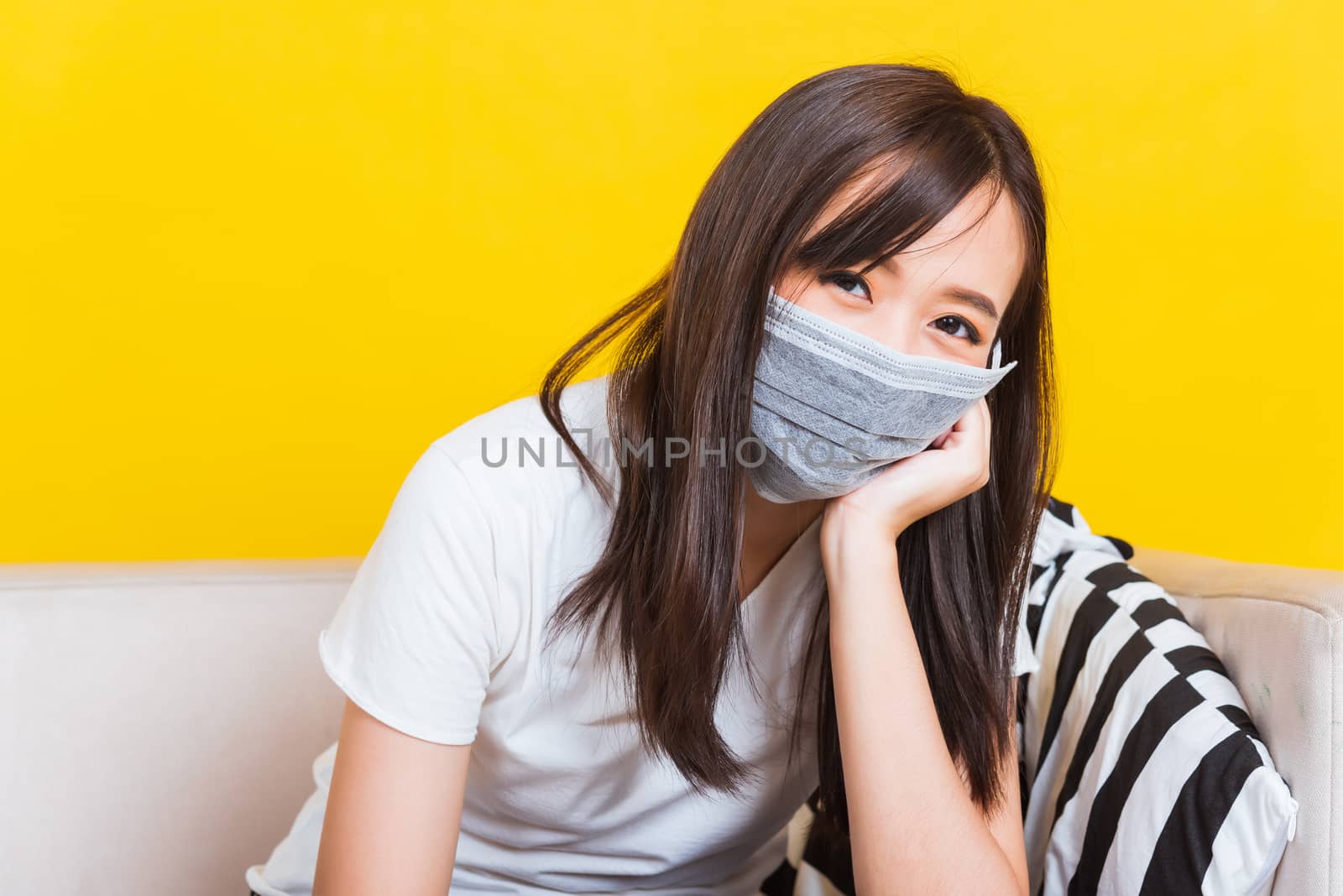 Portrait Asian of beautiful young woman sitting on sofa wearing medical face mask protective during Coronavirus studio shot isolated on yellow background