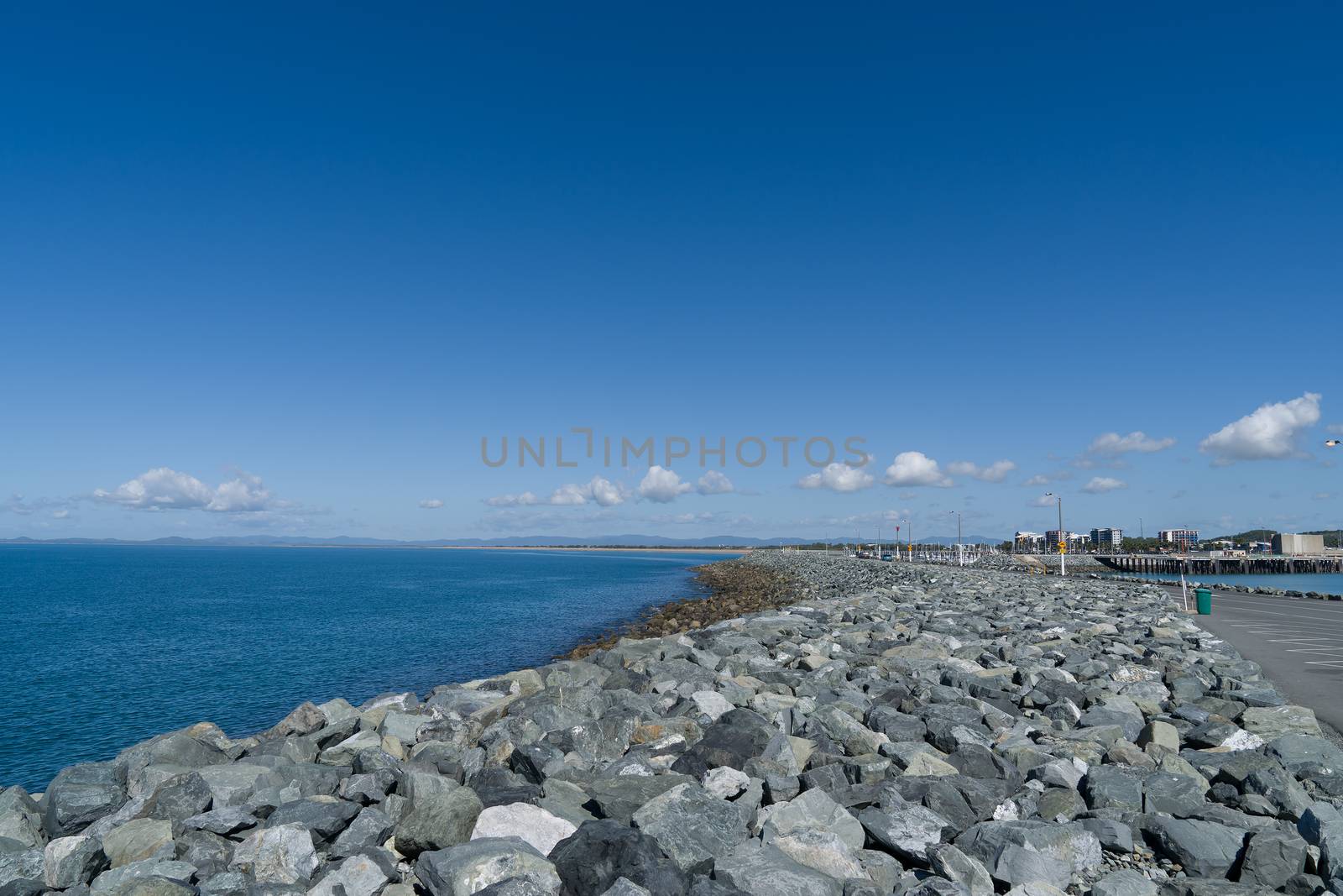 View from the rock wall of a marina breakwater out to the ocean and along the nearby beach, taking in some of the commercial and residential precinct behind