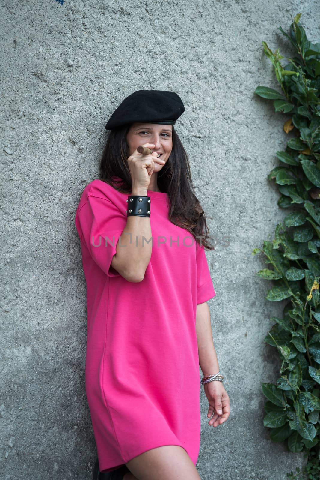 girl with dark hair smokes a big cigar wearing a black beret and a fuchsia dress and a bracelet, image of a woman posing in front of a light wall