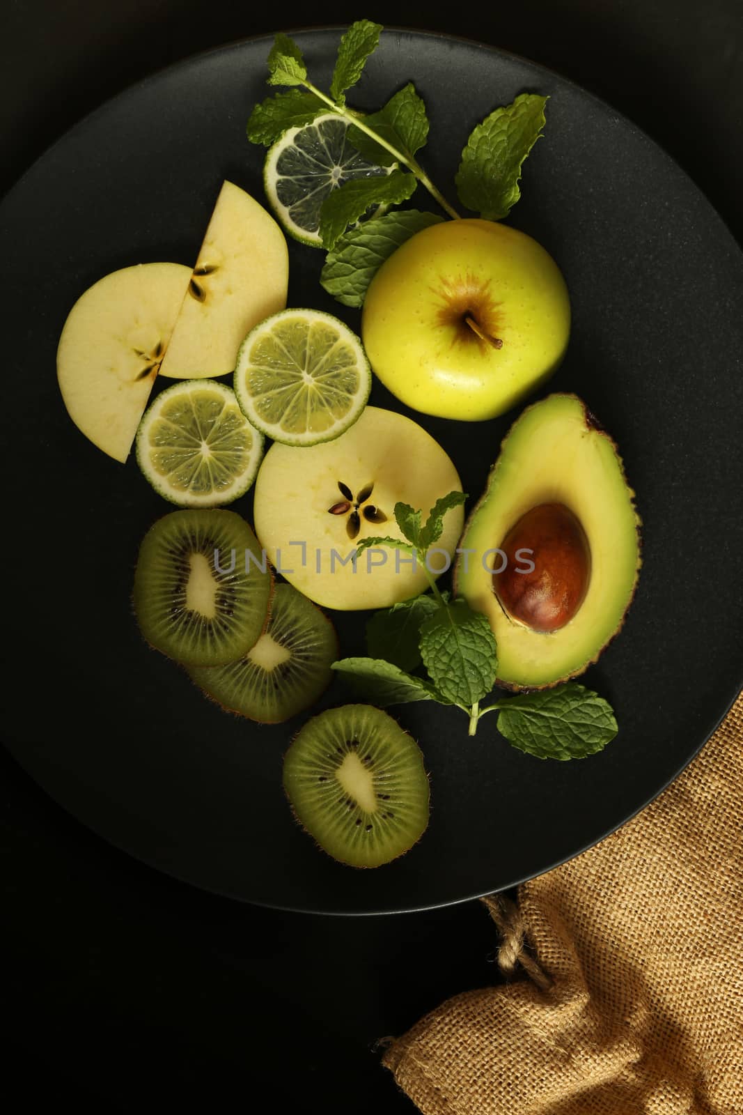 Multiple green fruits on round black plate on black background with canvas. Dramatic dark food.Flat lay, close up, top view