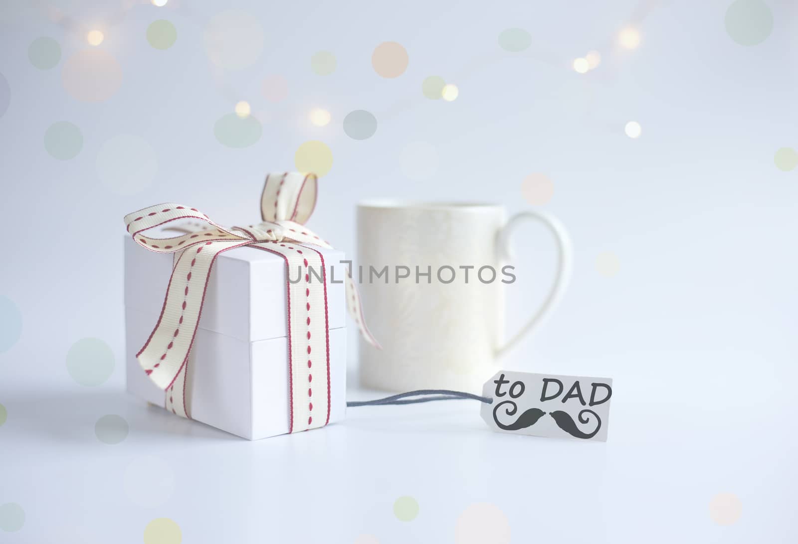 Cup of morning coffee, wrapped gift box, gift tag with word DAD and icon of moustache on light background with bokeh