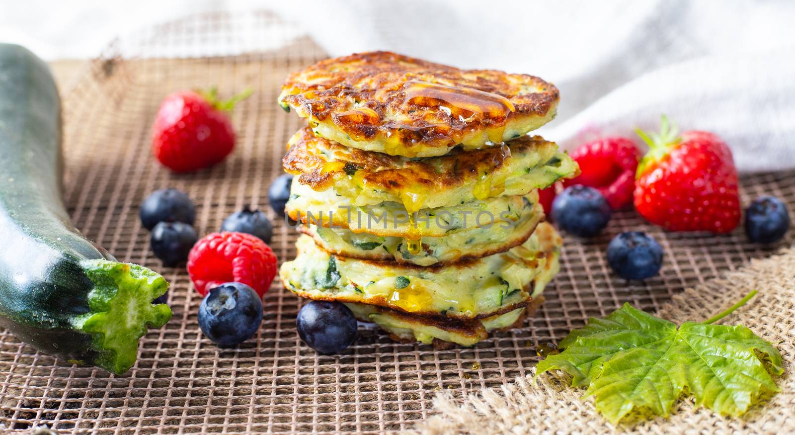 Fresh pile of delicious homemade courgette pancakes topped with honey maple syrup, summer fruits, zucchini and maple leaf on kitchen grill. Close up. Recipe, cooking, season, menu concept