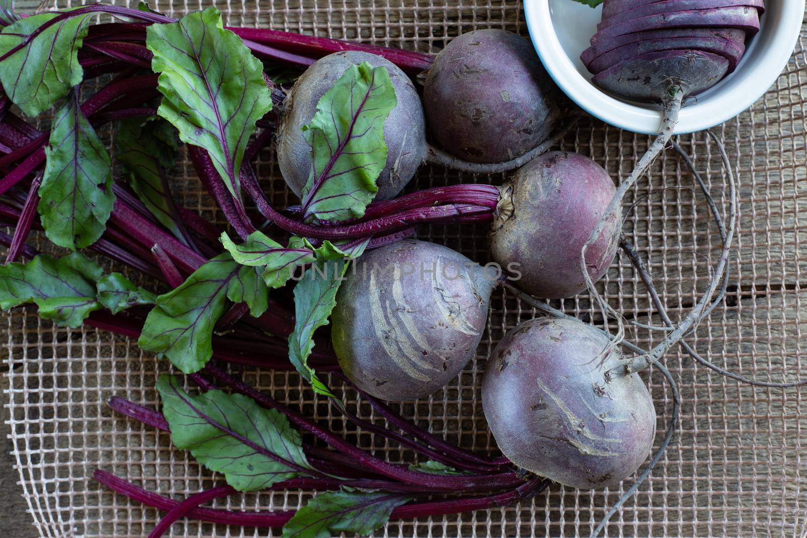 Bunch of fresh, spring, organic beetroot on kitchen grid, with slices of beetroot in ceramic plate on old wooden background. Top view. Rustic, dramatic, organic kitchen. Ingredients, menu. Healthy life concept