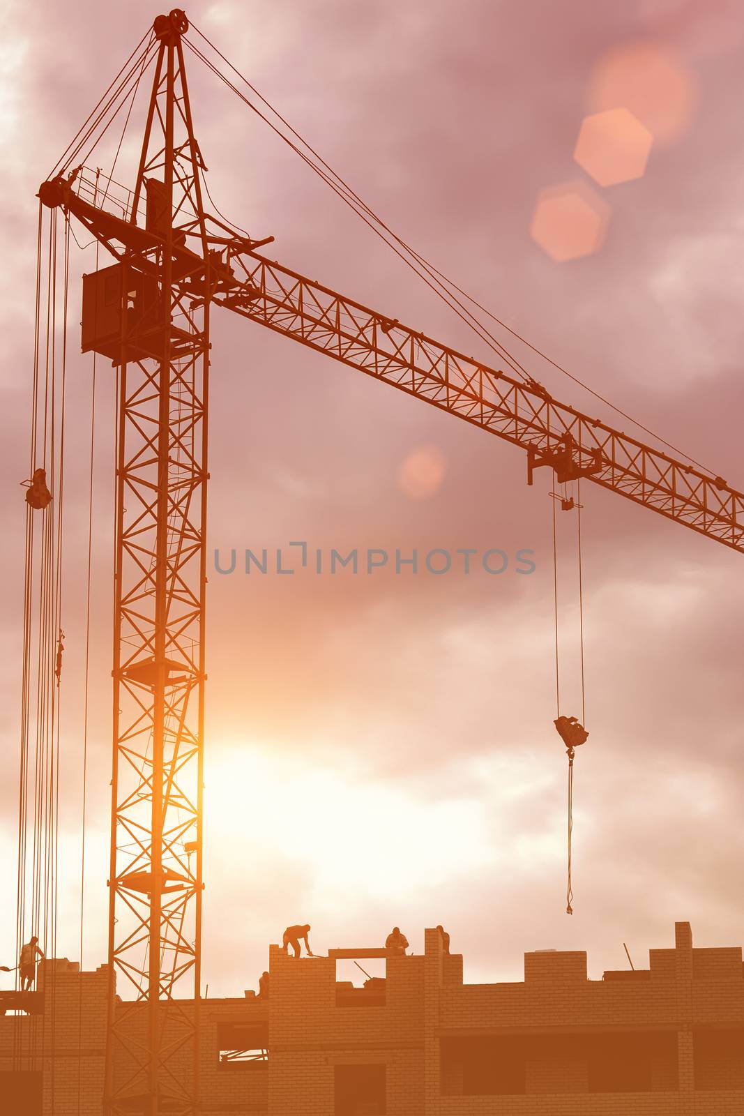 Silhouette of a team of construction workers in and a crane constructing a building on the background of the evening sunset sky.