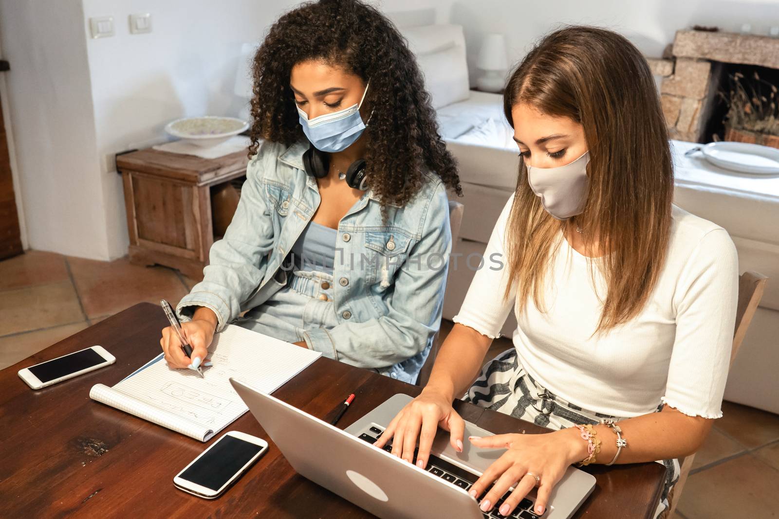 Lifestyle concept, work from home to reinvent your life: two young women, hispanic and caucasian, sitting on a table using a laptop with medical mask protection - New normal job activity