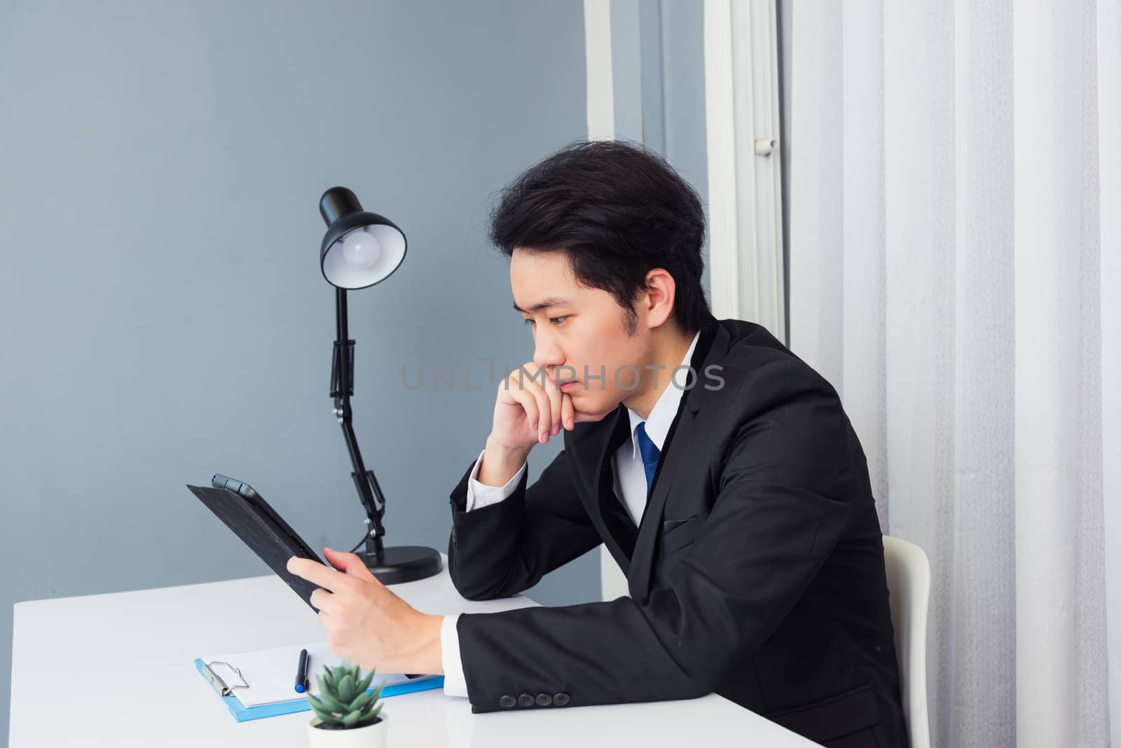 Work from home, Asian young businessman video conference call or facetime he smiling looking to tablet sitting on desk using smart tablet computer and thinking about work for profits at home office