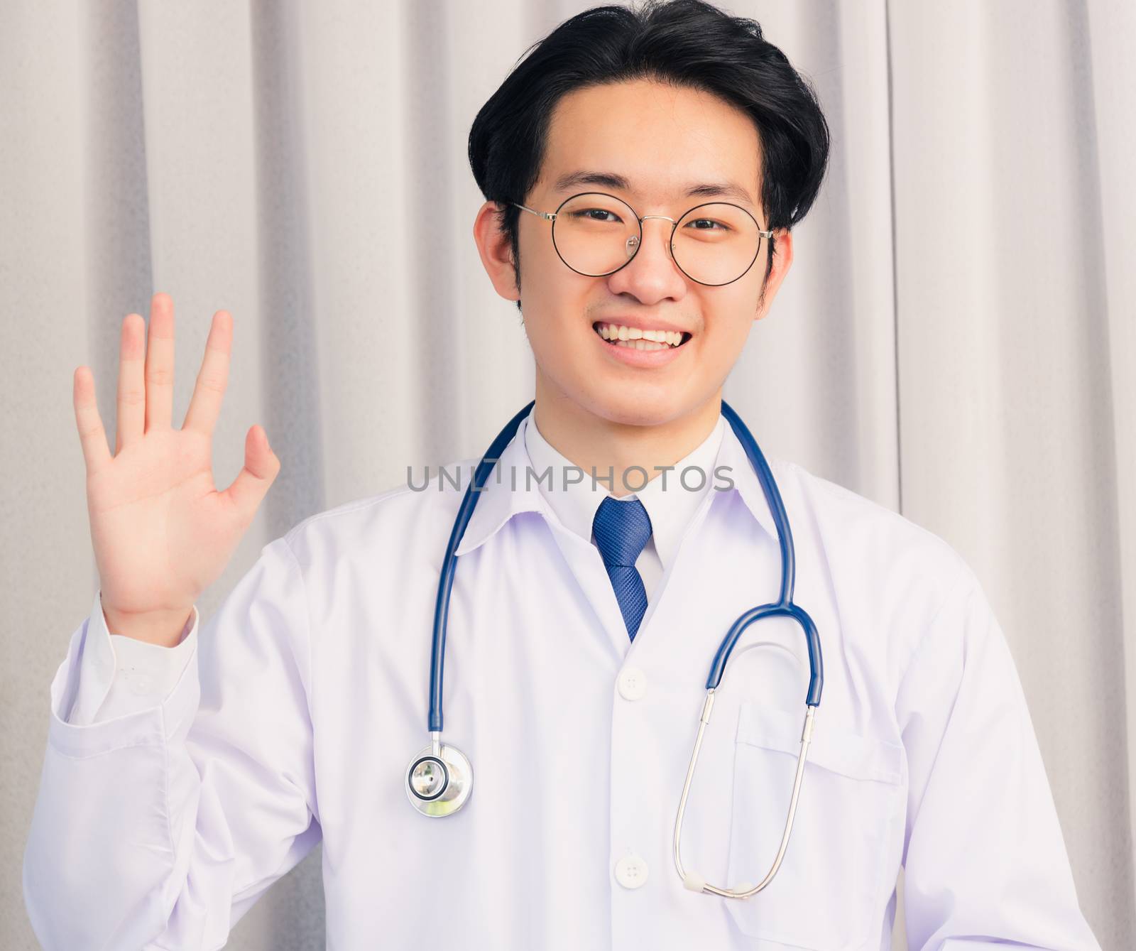 Portrait of Happy Asian young doctor handsome man smiling in uniform with stethoscope talking online video conference call or facetime raise his hand to say hello, healthcare medicine concept