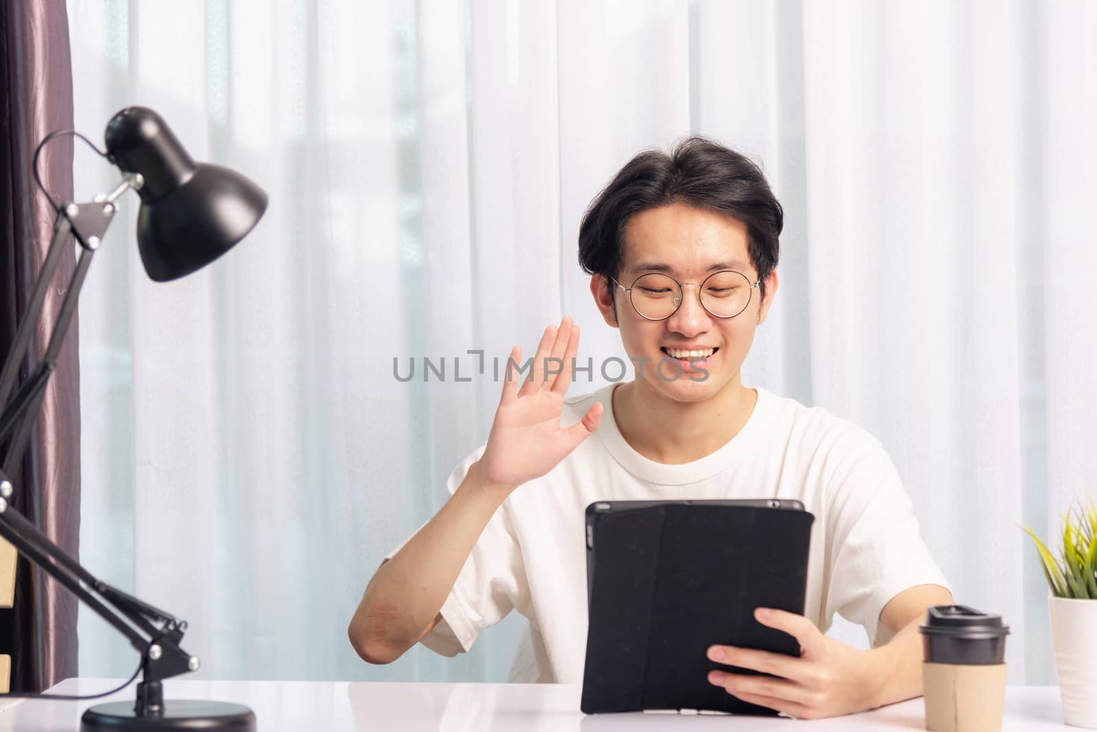 Happy Asian young business handsome man work from home office wear glasses, t-shirt comfortable he smiling and using a black modern smart digital tablet computer raise hand to say hello team on desk