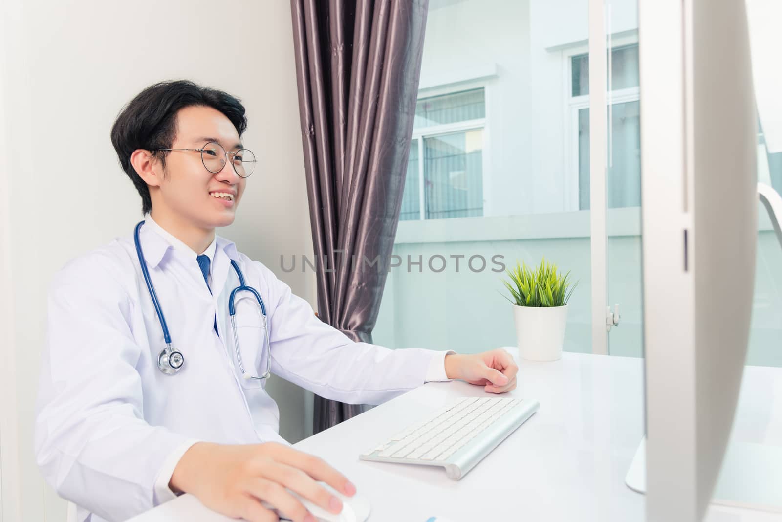 Asian young handsome doctor man wearing a doctor's dress and stethoscope video conference call or facetime with computer to patient he smiling sitting on desk at hospital office, Health medical care