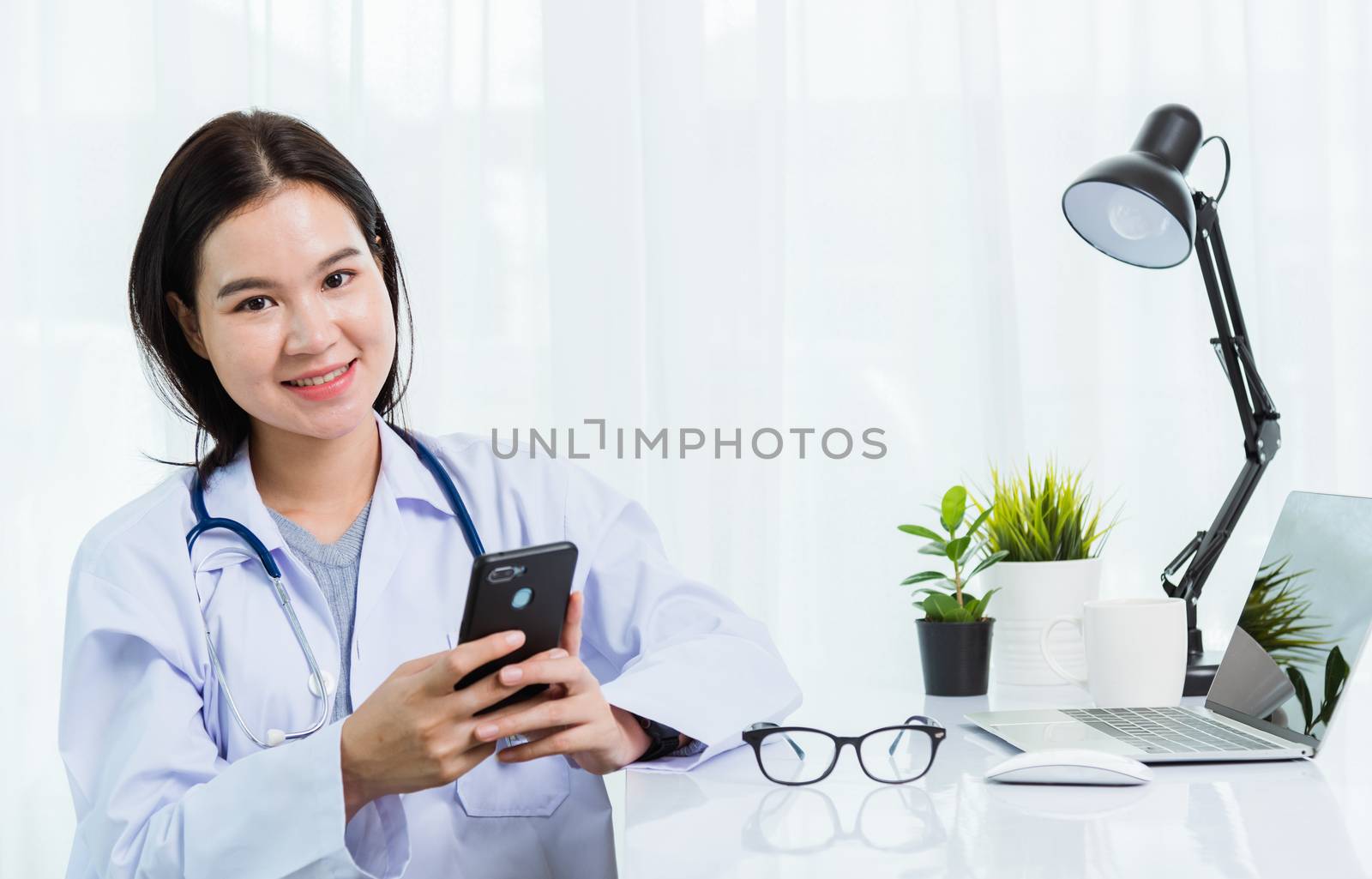 Asian doctor young beautiful woman smiling using working or holding with smart mobile phone and laptop computer at hospital desk office, technology healthcare medical concept