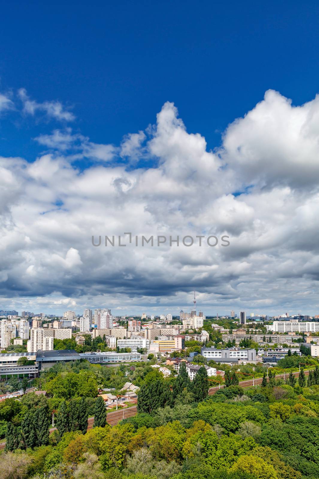 Cityscape of Kyiv in summer at noon with a green park, residential areas and a TV tower against a blue sky with thickening clouds. Bird's-eye view. Copy space.