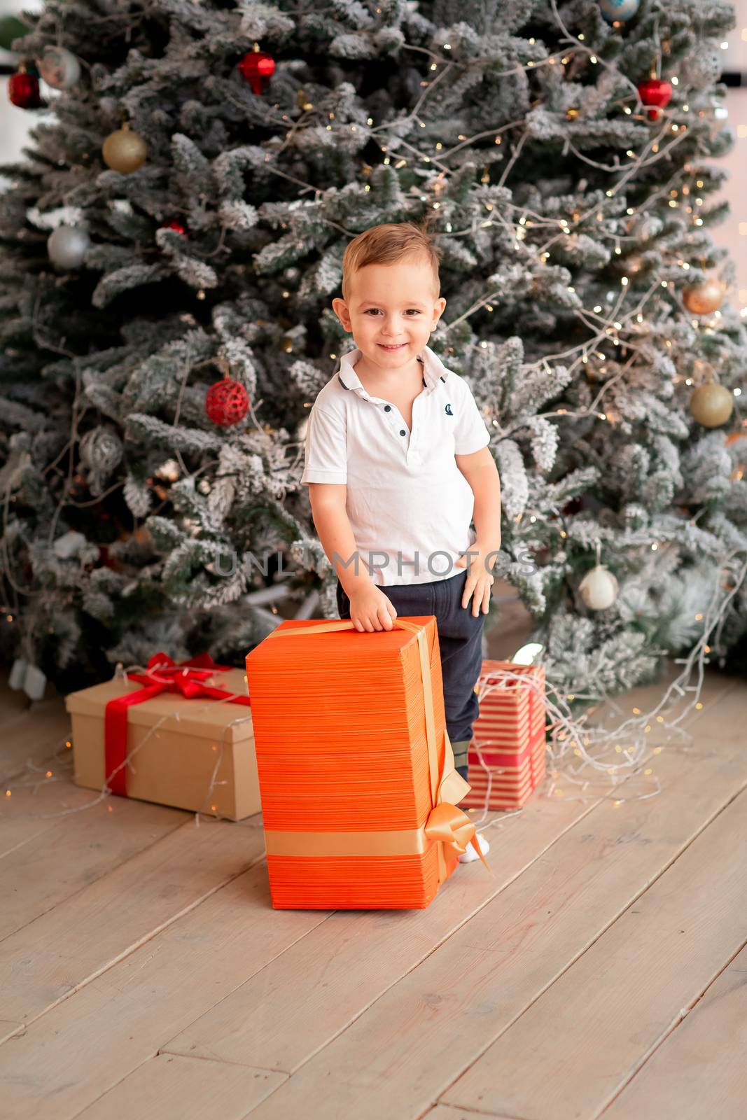 Cheerful cute baby boy with gifts. Kid holding big present and having fun near tree in the morning. Merry Christmas and Happy Holidays concept