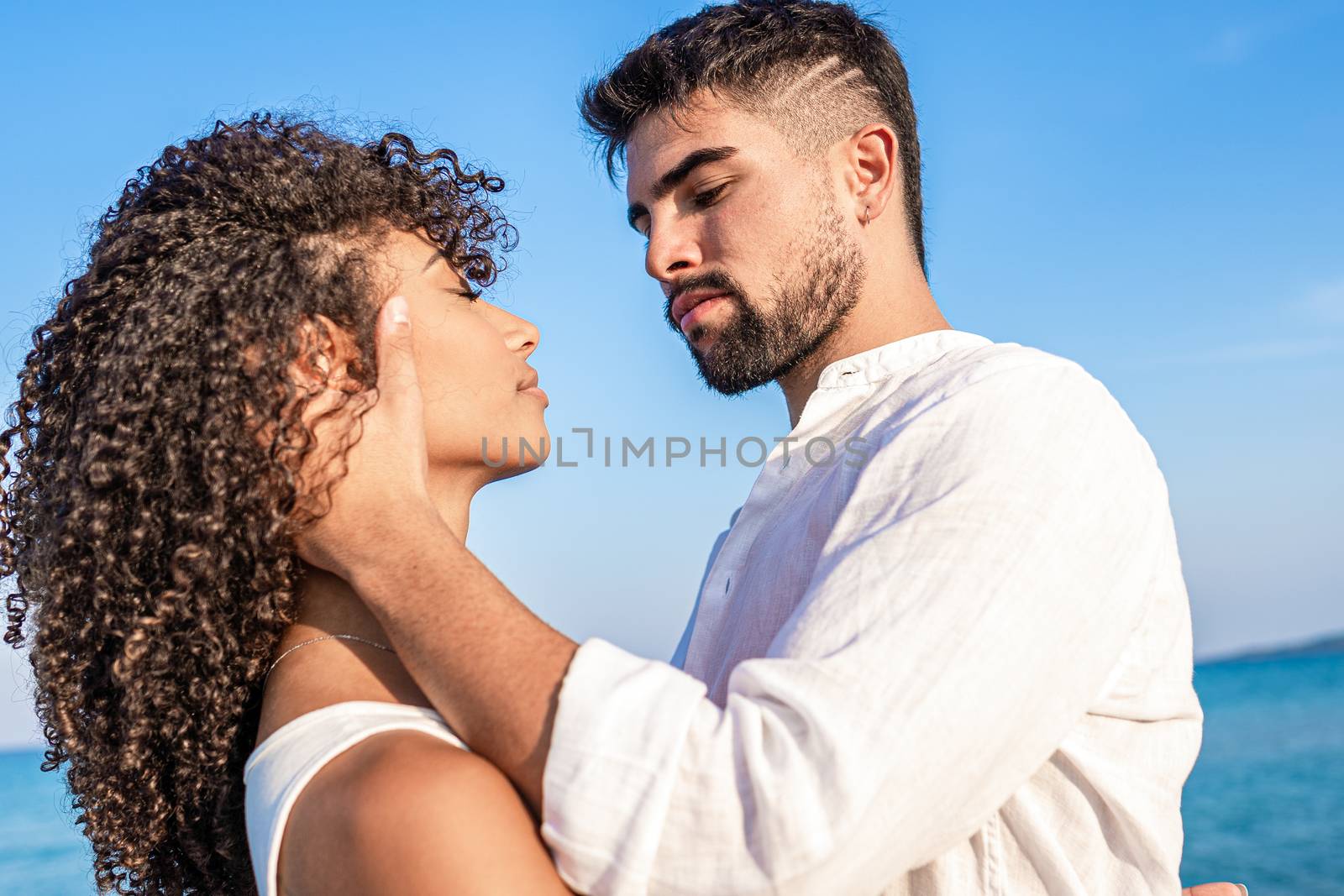 Young handsome bearded man in white shirt grabs his curly hispanic woman forcefully holding her head in his hands with serious expression - Bright and vivid color low angle view by robbyfontanesi