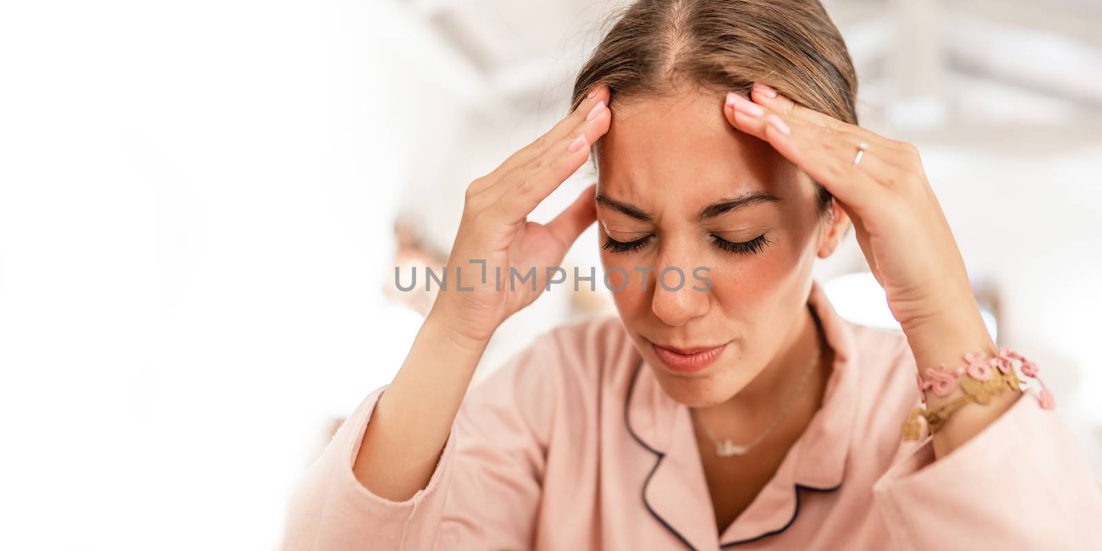 A young caucasian woman wearing pink pajama holds her head in her hands and grimaces in pain from the migraine headache