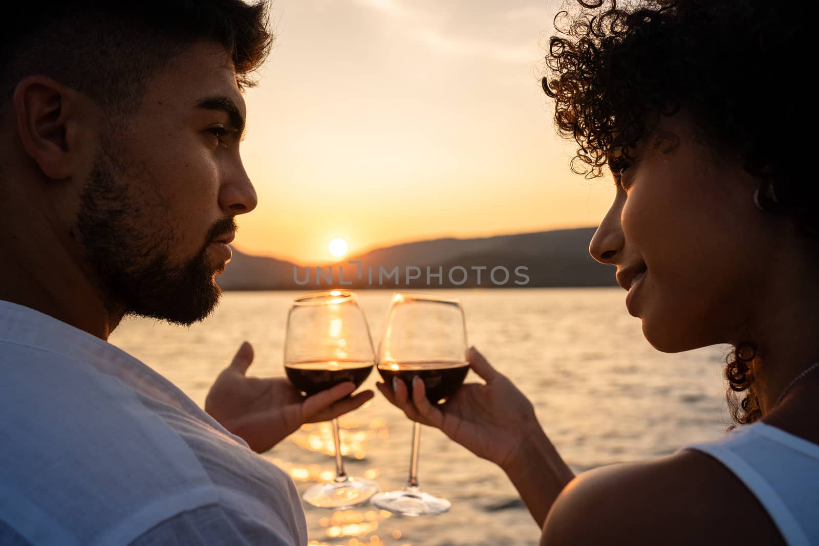 Romantic close-up of a mixed race couple toast with glasses of red wine crossed by the light of the setting sun on a lake by robbyfontanesi