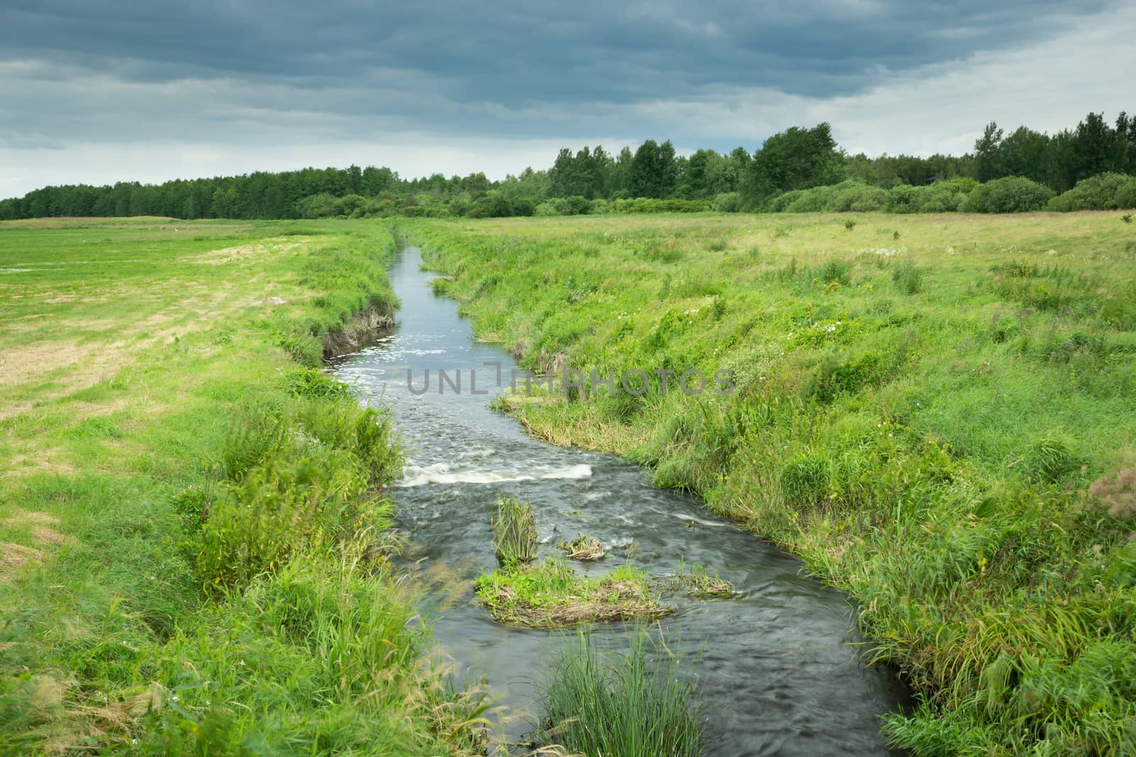 A small river flowing through meadows and clouds in the sky, summer view