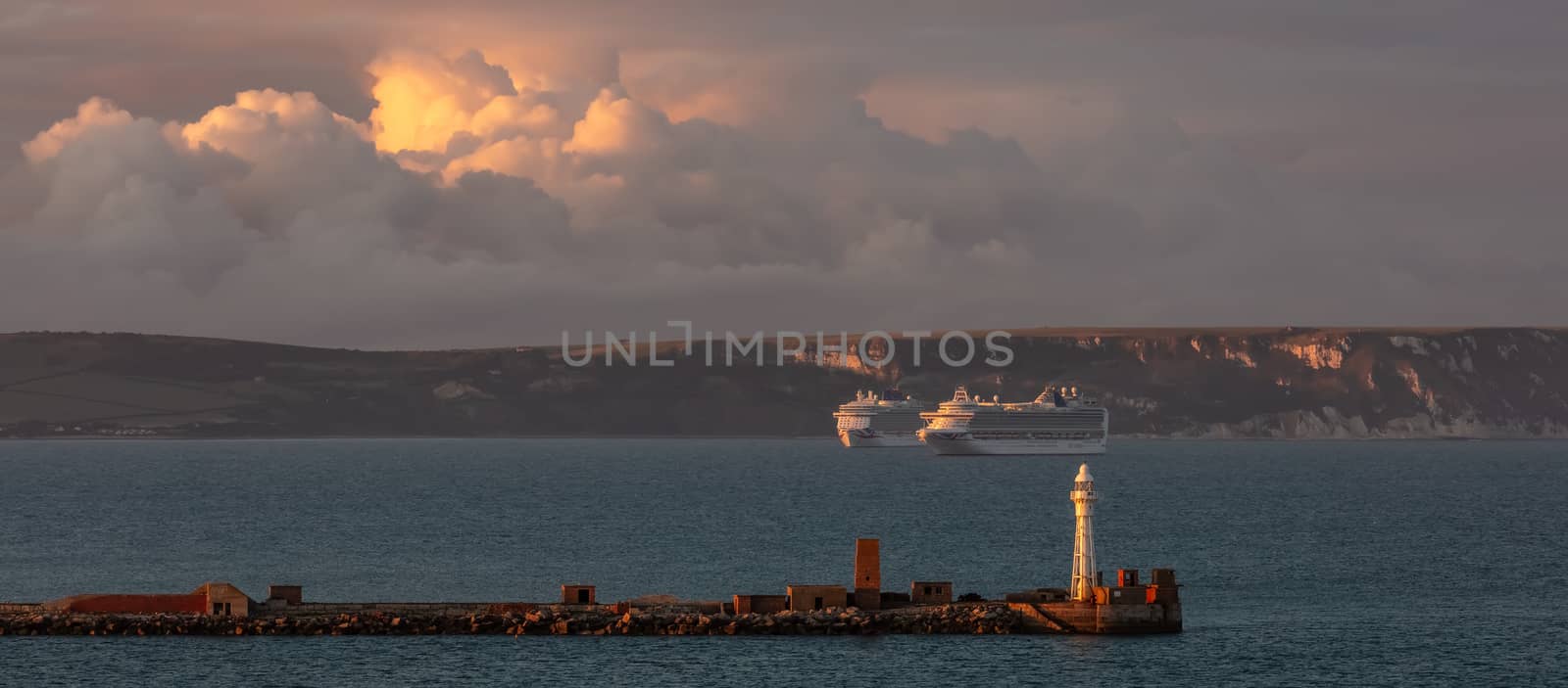 Portland, United Kingdom - July 1, 2020: Beautiful panoramic shot of Portland harbour lighthouse with two P&O cruiseships anchored in the distance in Weymouth Bay. Beautiful sunset clouds.