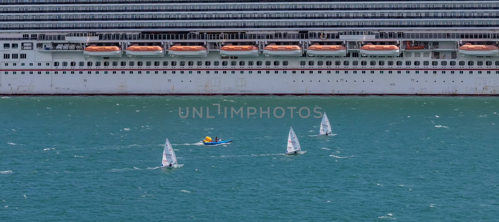 Portland harbour, United Kingdom - July 2, 2020: High Angle aerial panoramic shot of the laser class sailing racing dinghies and a rescue boat sailing by a huge cruise ship in Portland harbour.