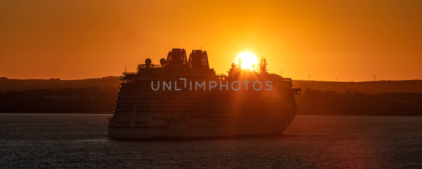 Weymouth Bay, United Kingdom - July 10, 2020: Amazing panoramic shot of P&O cruise ship Britannia anchored in Weymouth Bay at sunset. Sun setting down right above the ship casting orange color on it