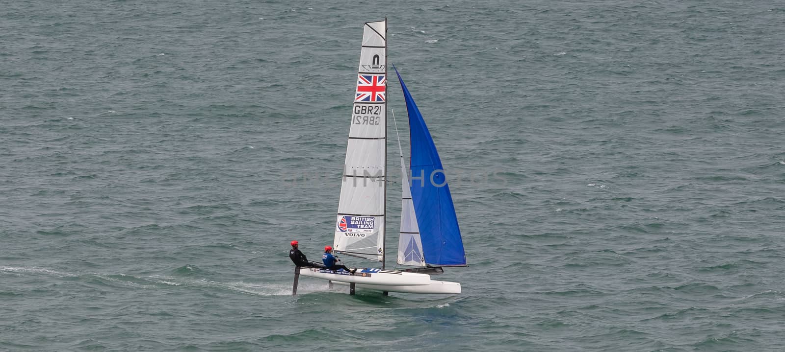 Portland harbour, United Kingdom - July 1, 2020: High Angle aerial panoramic shot of racing catamaran of the British Sailing Team. Two sailors on it wearing red helmets, British flag on the sails.