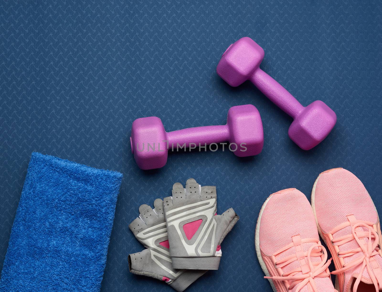 pair of purple dumbbells, sports gloves and pink gym shoes on a blue neoprene mat, top view