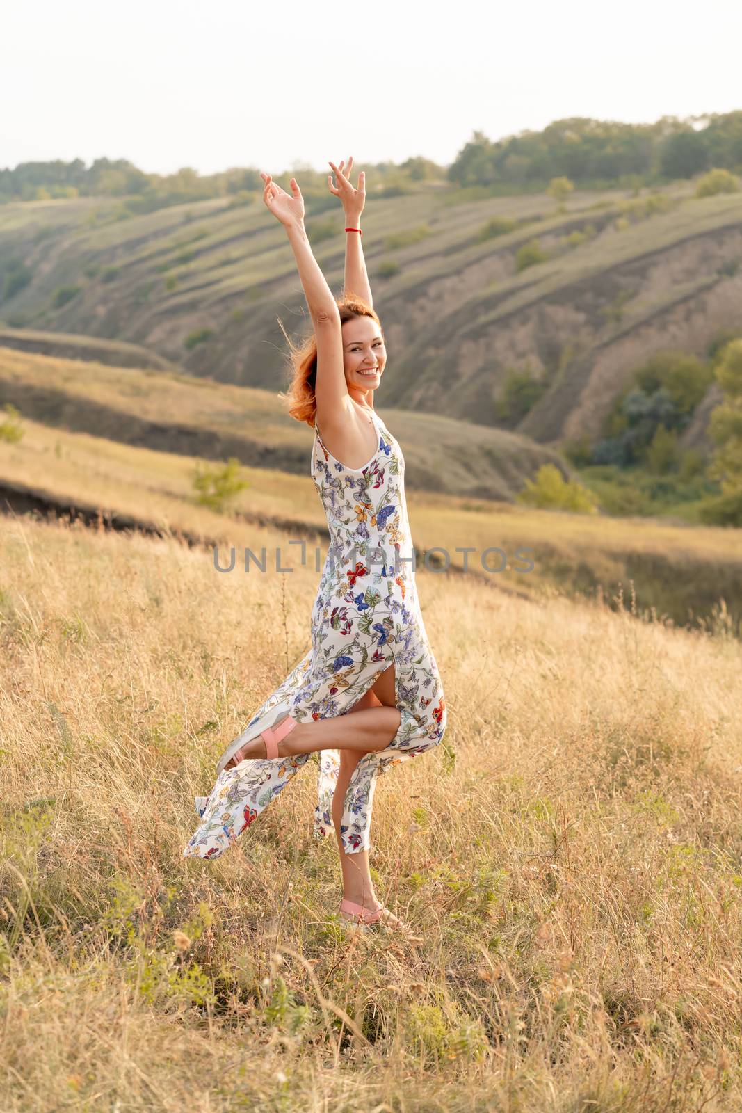 Beautiful red-haired girl is having fun and dancing in a field at sunset.