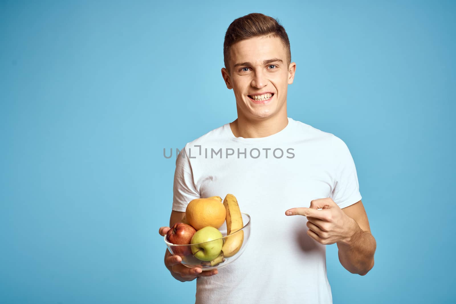 Happy man with fresh fruits gesturing with hands blue background white t-shirt vitamins bananas oranges apples by SHOTPRIME