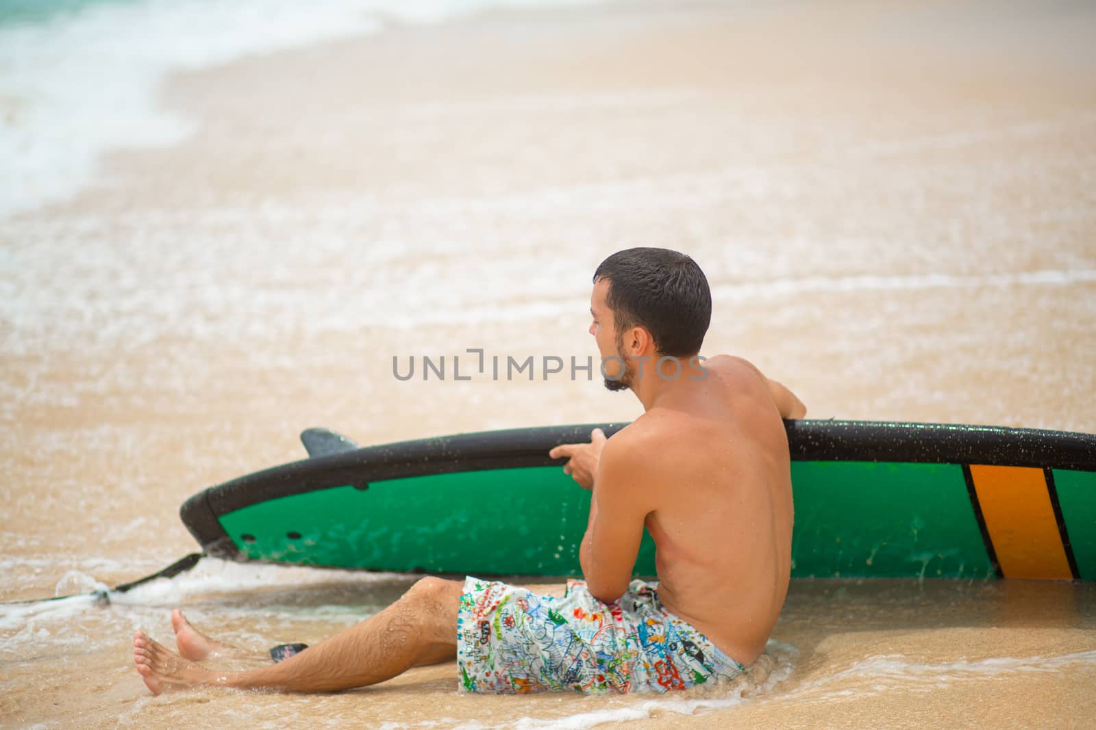 The guy is resting on a sandy tropical beach, after riding a surf. Healthy active lifestyle in summer vocation.