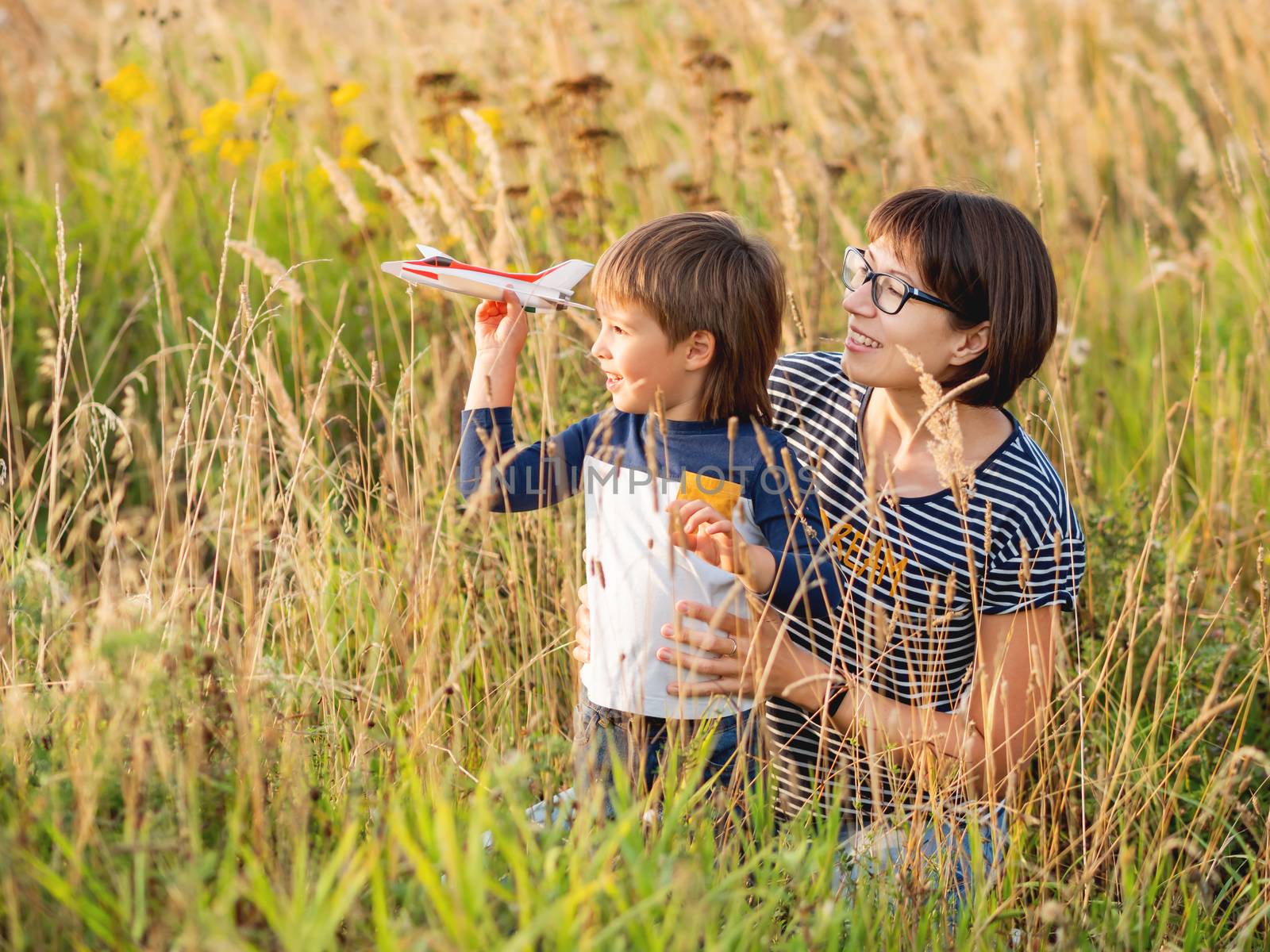 Cute boy and his mother play with toy airplane. Happy kid dreams to be a pilot. Boy is planning for the future. Mom and son on field at golden sunset hour at autumn season.