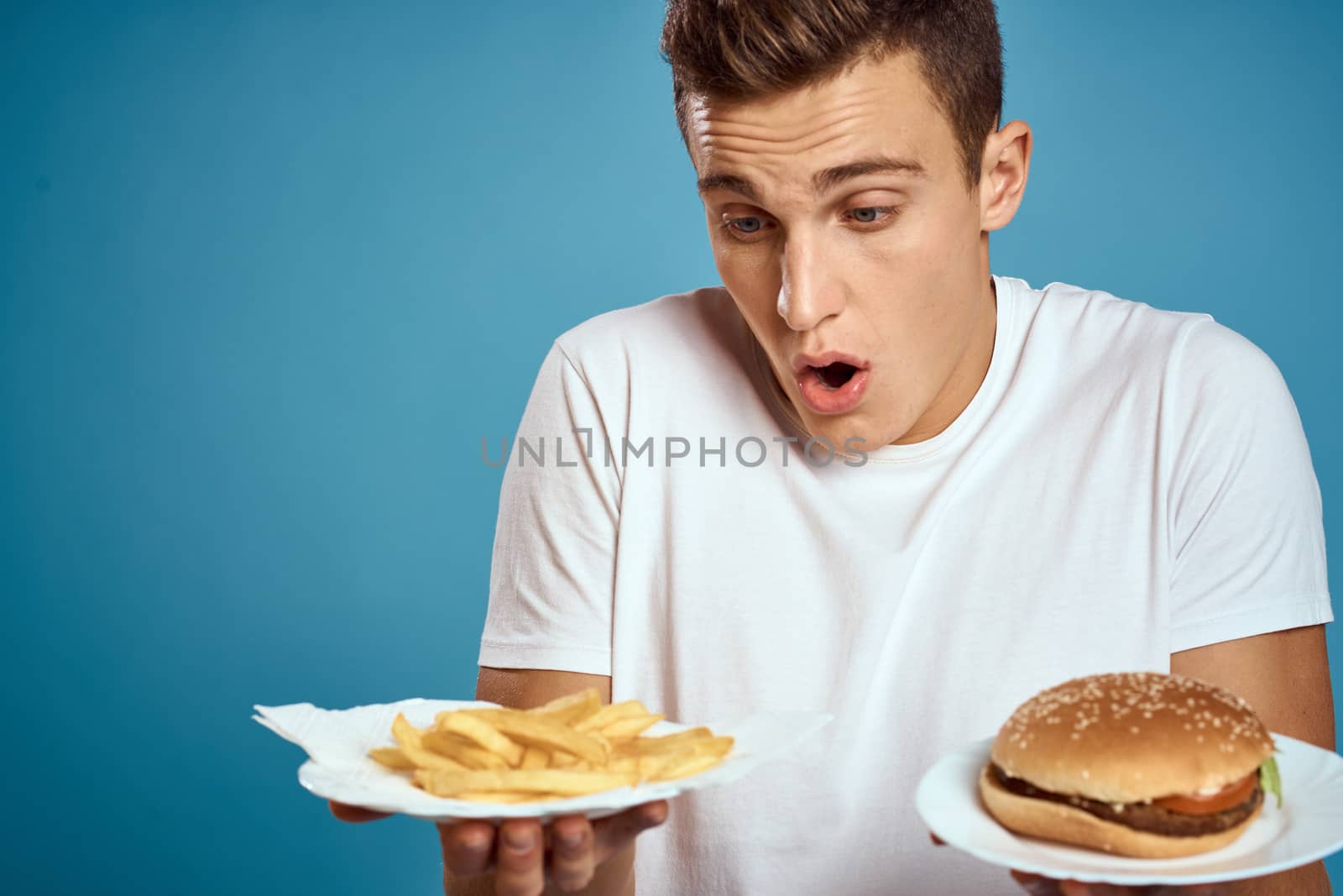 young guy with fries and hamburger on blue background interested look emotions fast food calories cropped view Copy Space. High quality photo