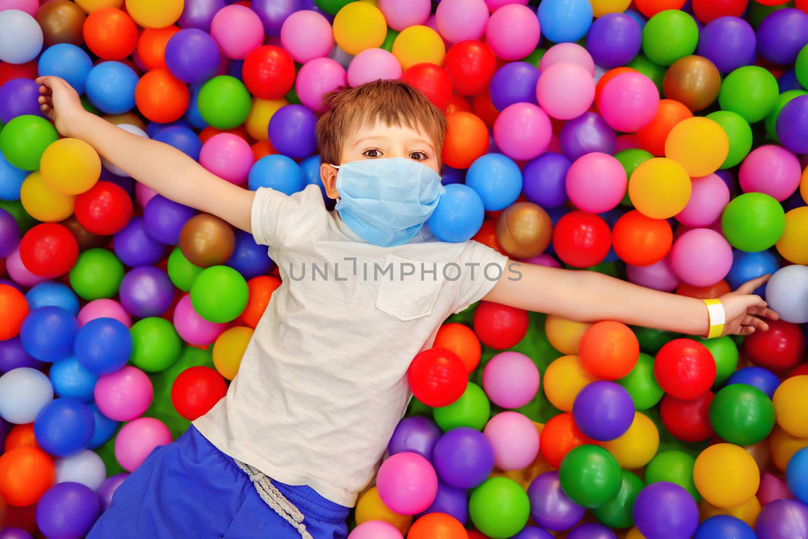 Boy in protective facemask at entertainment center. Joyful kid having fun among colorful soft balls. Kid in facemask lying in dry pool with colorful soft balls. Birthday party during covid coronavirus by synel