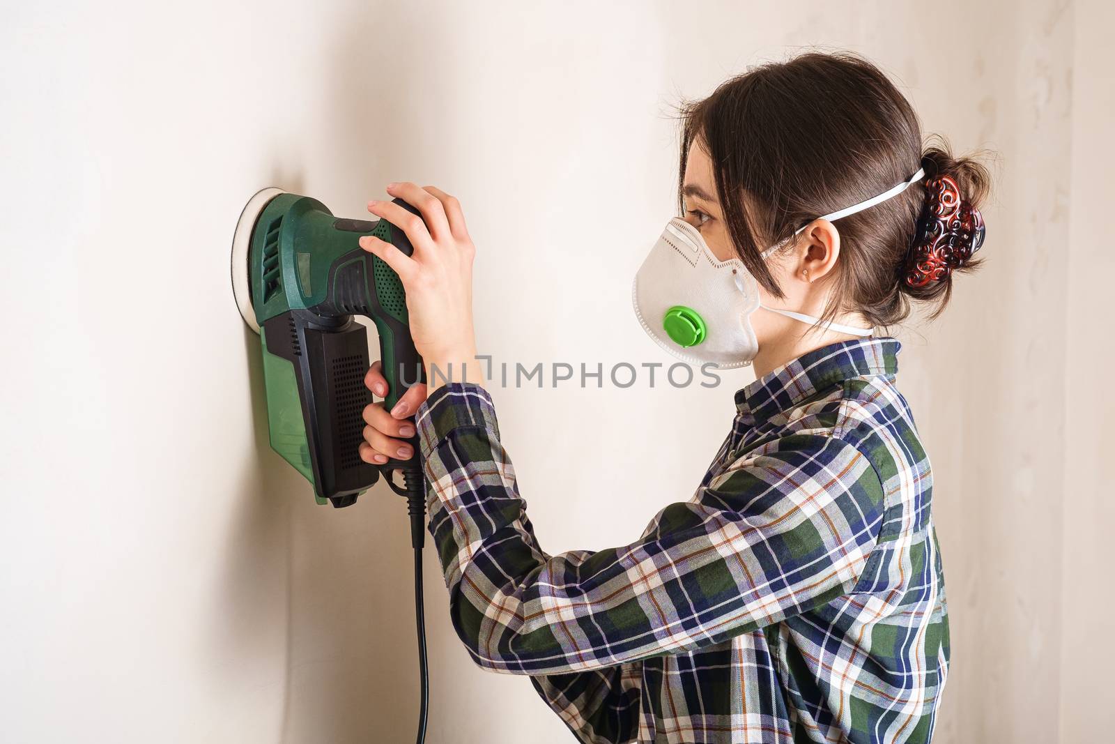 Woman in protective mask working with electric sander to smooth plaster wall surface by Frostroomhead