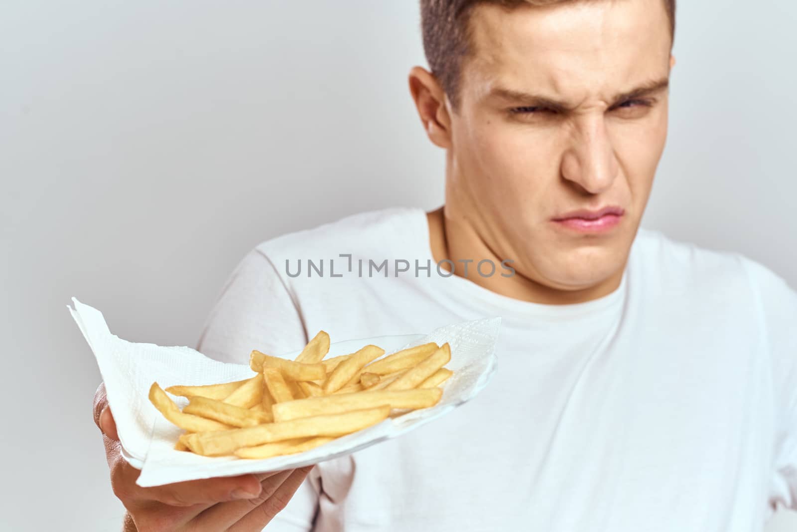 young guy with french fries in a box and in a white t-shirt on a light background cropped view. High quality photo