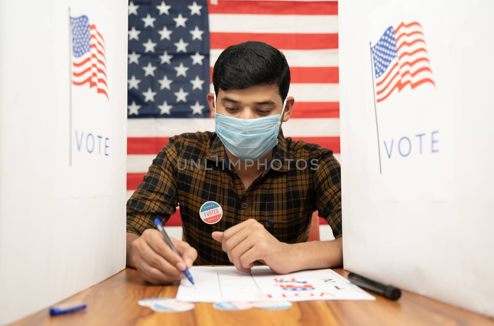 Young man in medical mask busy inside the polling booth with US flag as background - Concept of in person voting with covid-19 safety measure at US election. by lakshmiprasad.maski@gmai.com