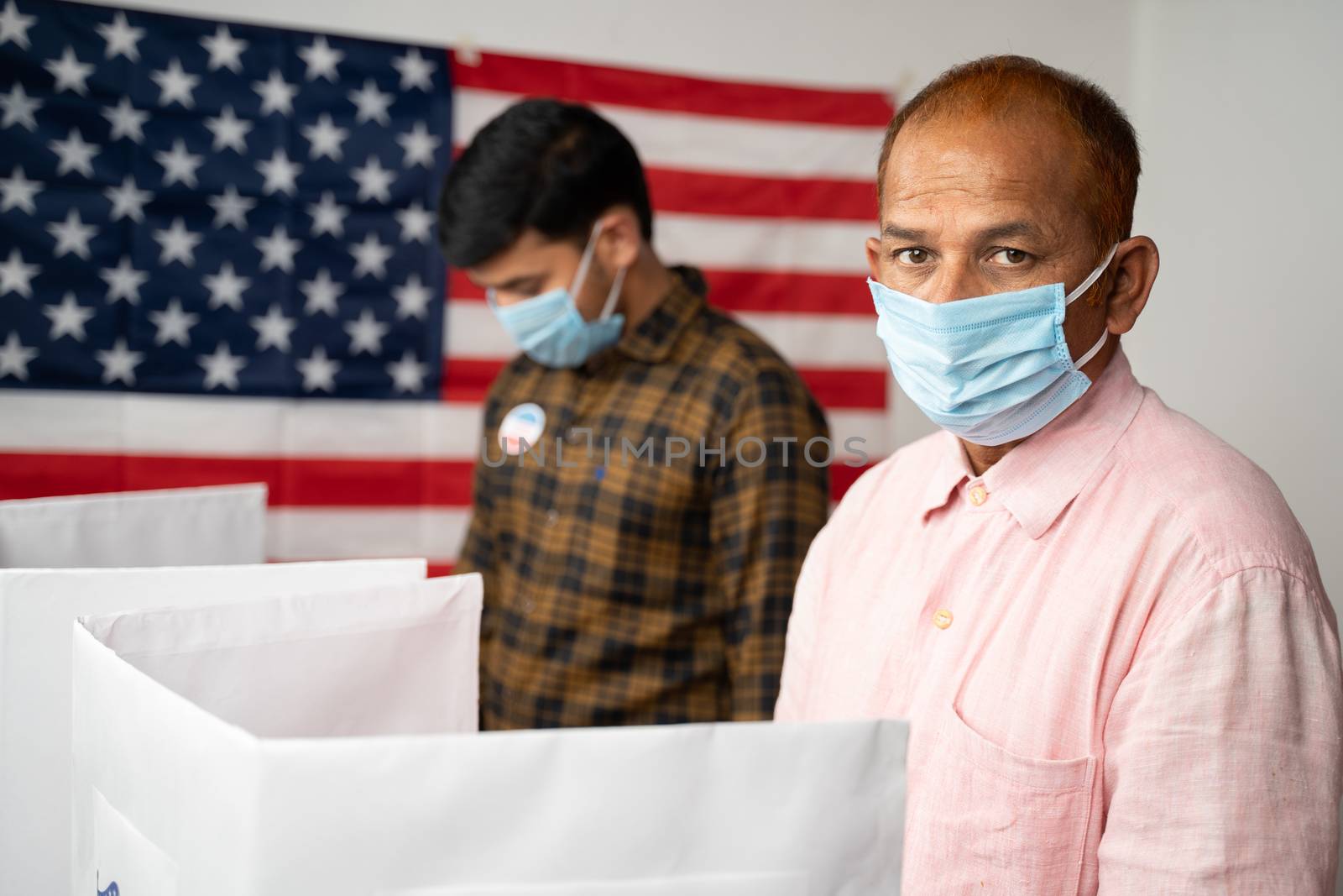 People with mask busy at polling booth for voting with US flag as background - concept of in person voting with covid-19 safety measures at us election.