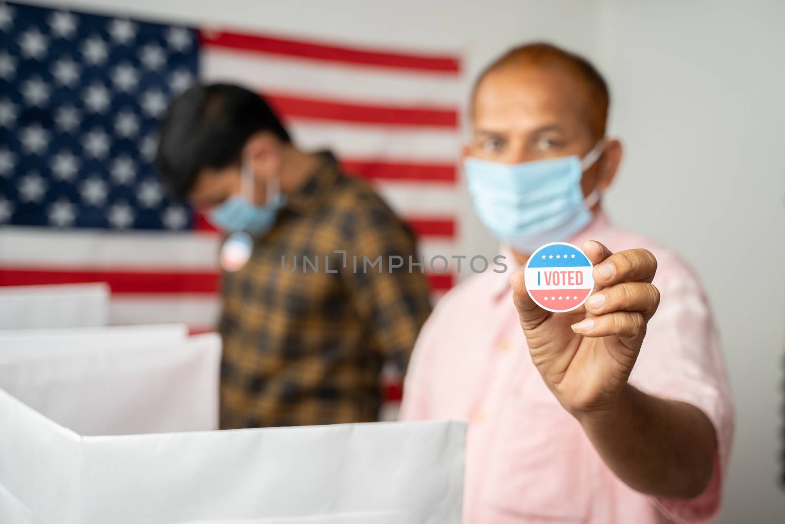 Man in medical mask showing I voted Sticker at polling booth with US flag as background - concept in person voting at US election. by lakshmiprasad.maski@gmai.com