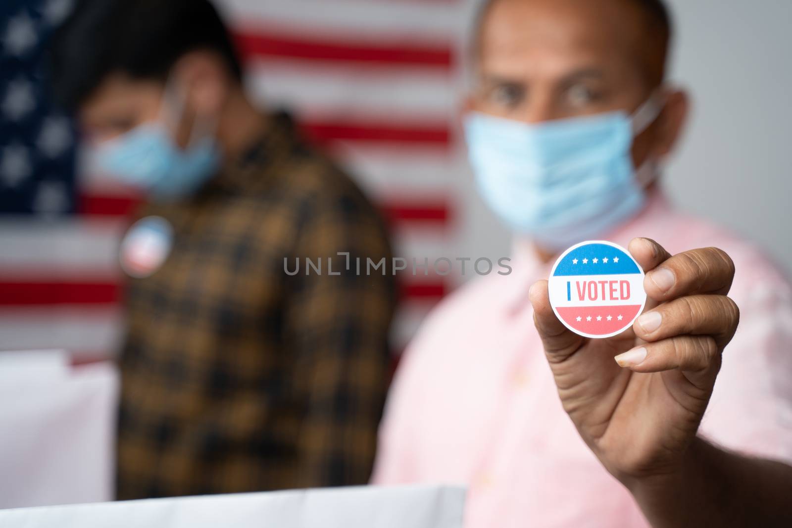 Close up of Hands, Man in medical mask showing I voted Sticker at polling booth with US flag as background - concept in person voting at US election. by lakshmiprasad.maski@gmai.com