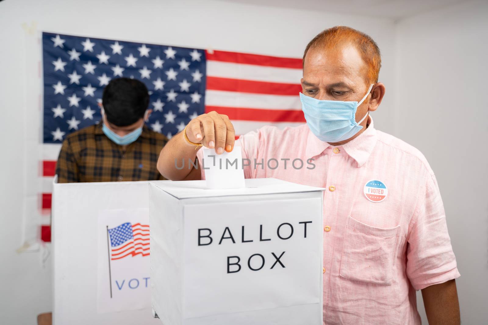Man in medical mask placing ballot paper inside the ballot box - Concept of in person voting and people busy at polling booth at US Election. by lakshmiprasad.maski@gmai.com
