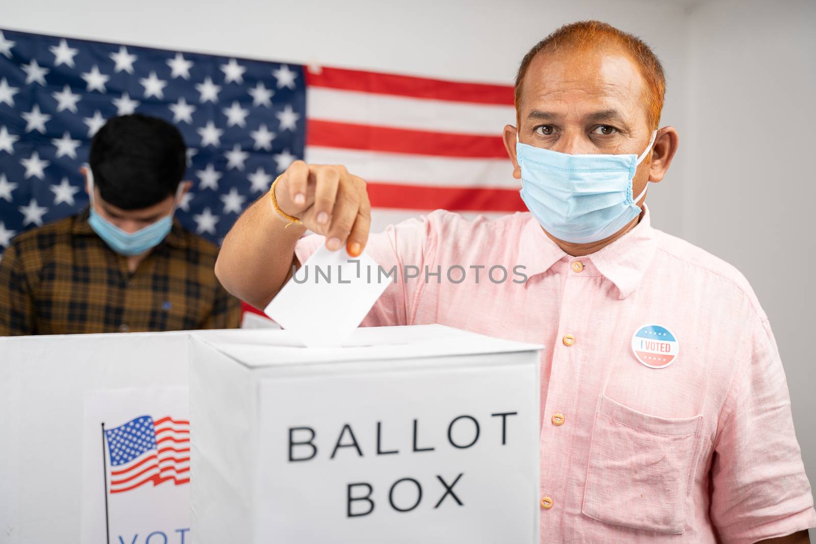 Man in medical mask placing ballot paper inside the ballot box while looking at camera - Concept of in person voting and people busy at polling booth at US Election