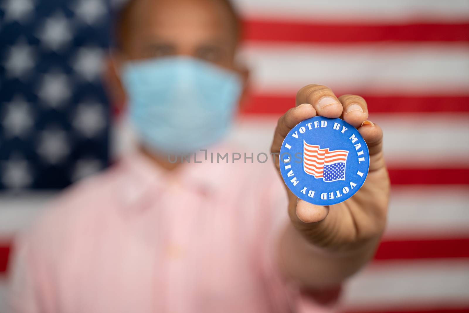 Man in medical mask showing I voted by mail sticker with US flag as background - Concept of mail in voting at USA election. by lakshmiprasad.maski@gmai.com