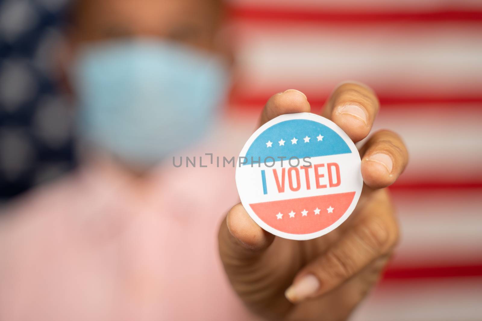 Close up shot man in medical mask showing I voted Sticker and putting on shirt with US flag as background - concept of US election