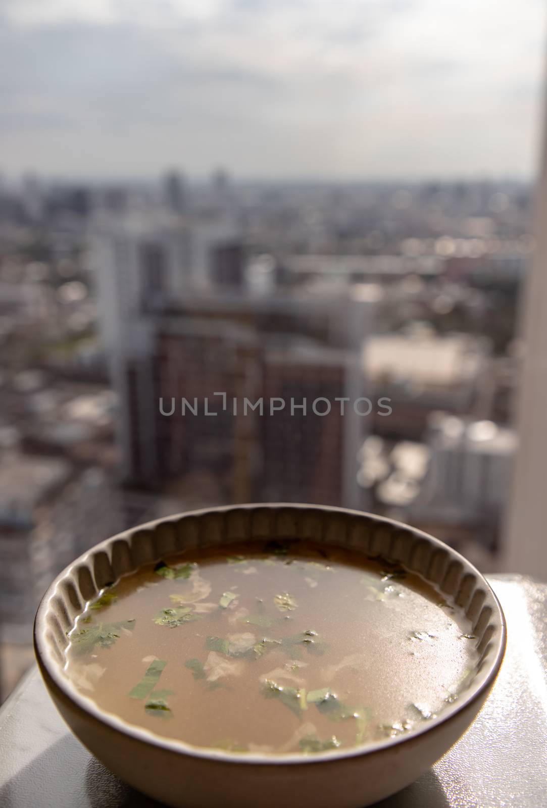Chicken broth with coriander in white bowl over marble table with city view at morning. Selective focus.