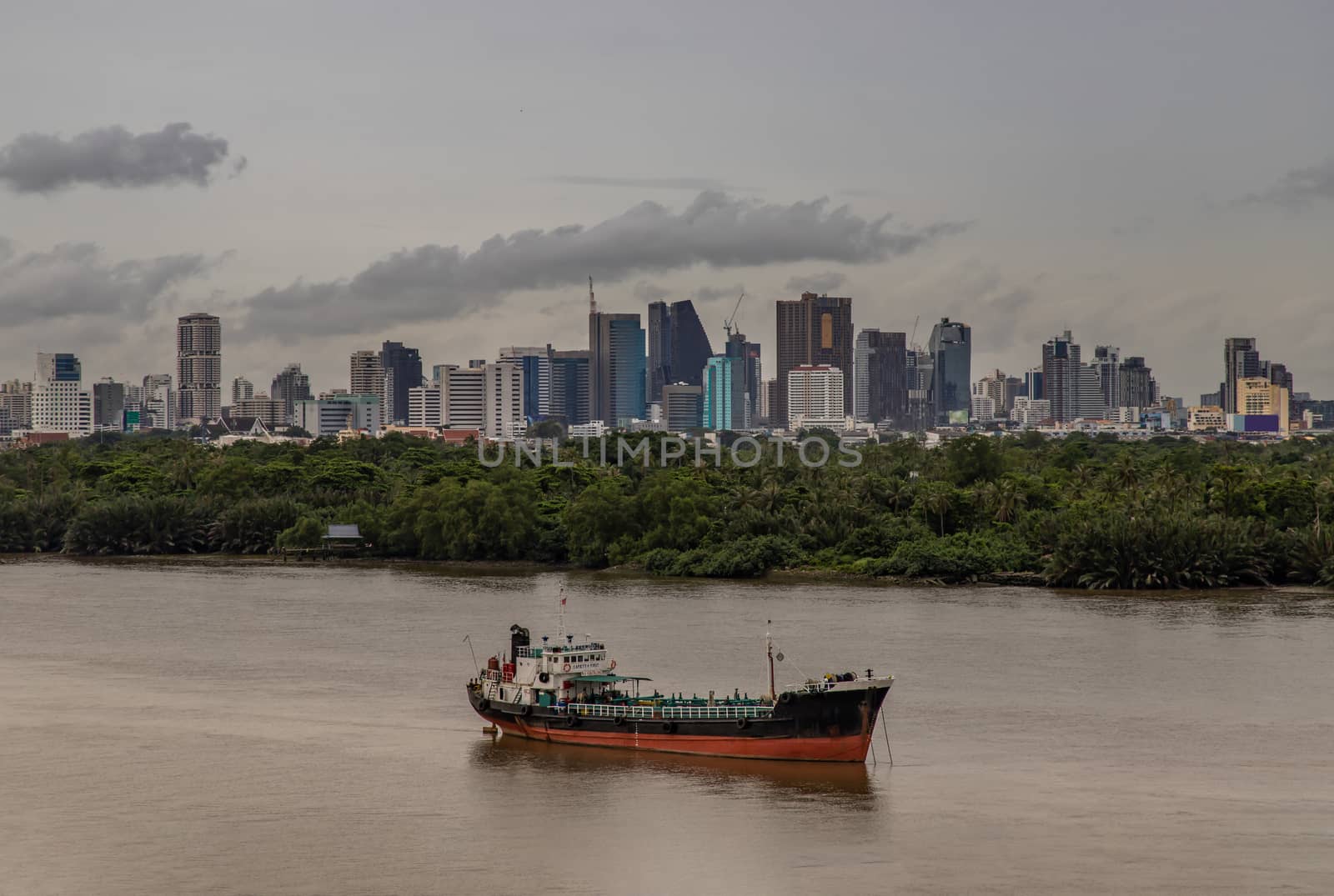 Bangkok, thailand - Sep 20, 2020 : A cargo ship parked in the middle of the river and in front of a green forest with skyscrapers at evening by the Chao Phraya River.