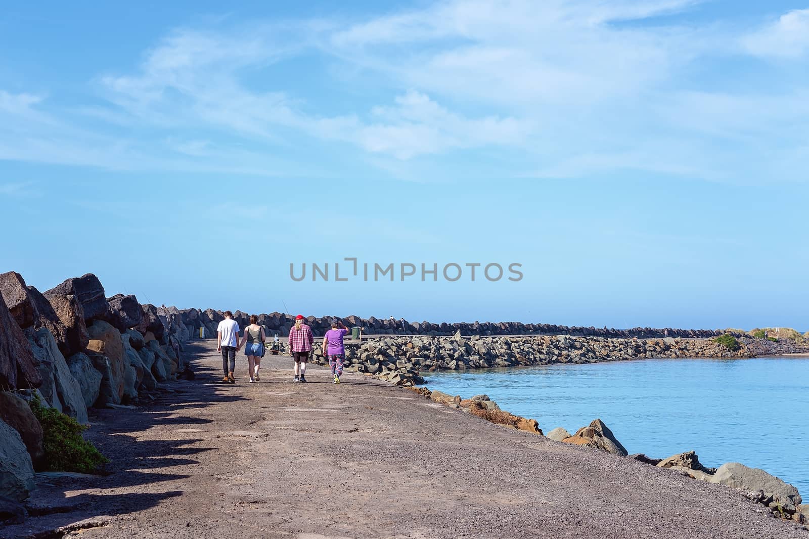 A family group of two young people and two older persons, unidentified, walking slowing along a marina breakwater wall on a bright sunny day with blue sky and water.