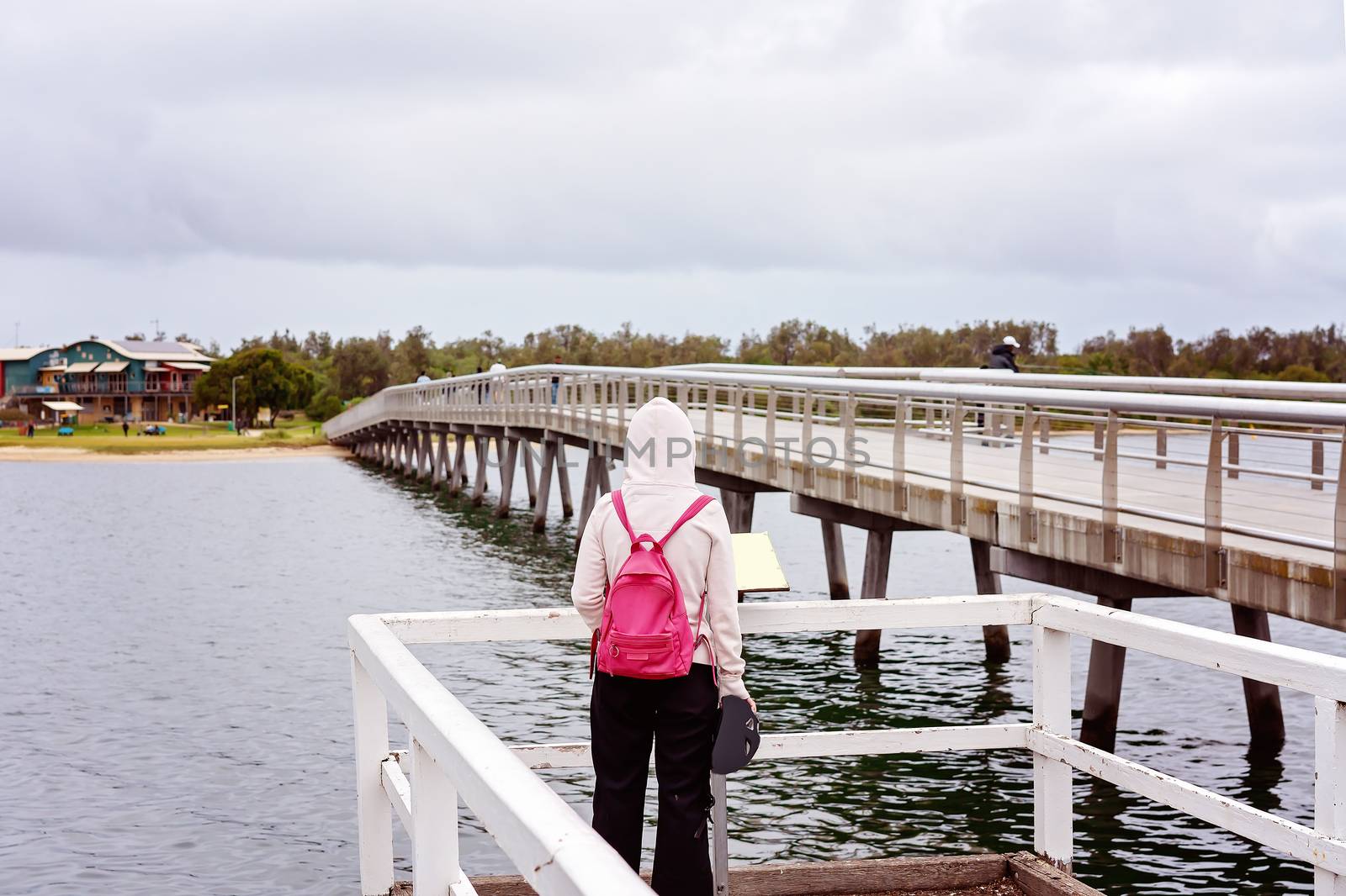 A young person in a pink coat and hood with a pink backpack standing reading a sign on a jetty beside a river