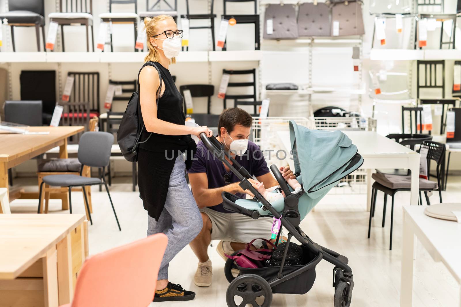 Young couple with newborn in stroller shopping at retail furniture and home accessories store wearing protective medical face mask to prevent spreading of corona virus when shops reopen. by kasto