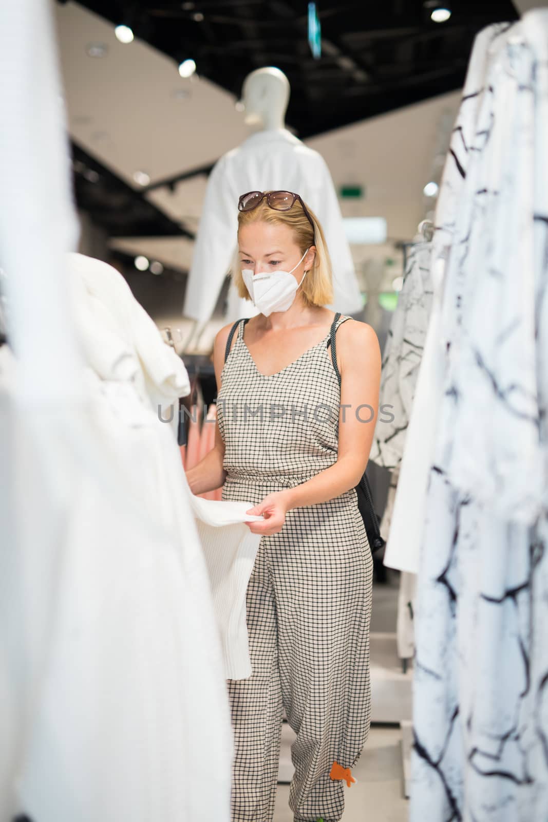 Fashionable woman wearing protective face mask shopping clothes in reopen retail shopping store. New normal lifestyle during corona virus pandemic by kasto