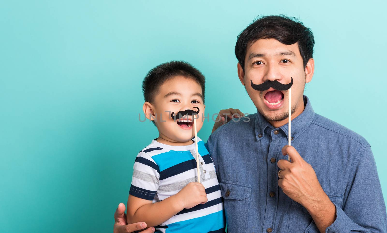 Family funny happy hipster father and his son kid holding black mustache props for the photo booth close face, studio shot isolated on a blue background, November men health awareness